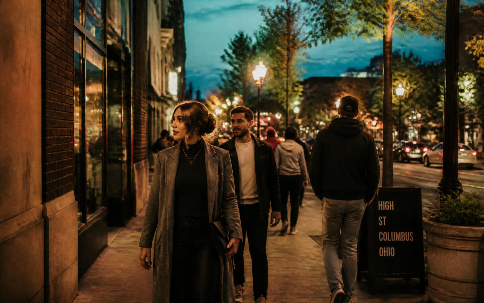 People in layered fall jackets walking a busy High Street sidewalk in Columbus, Ohio at dusk